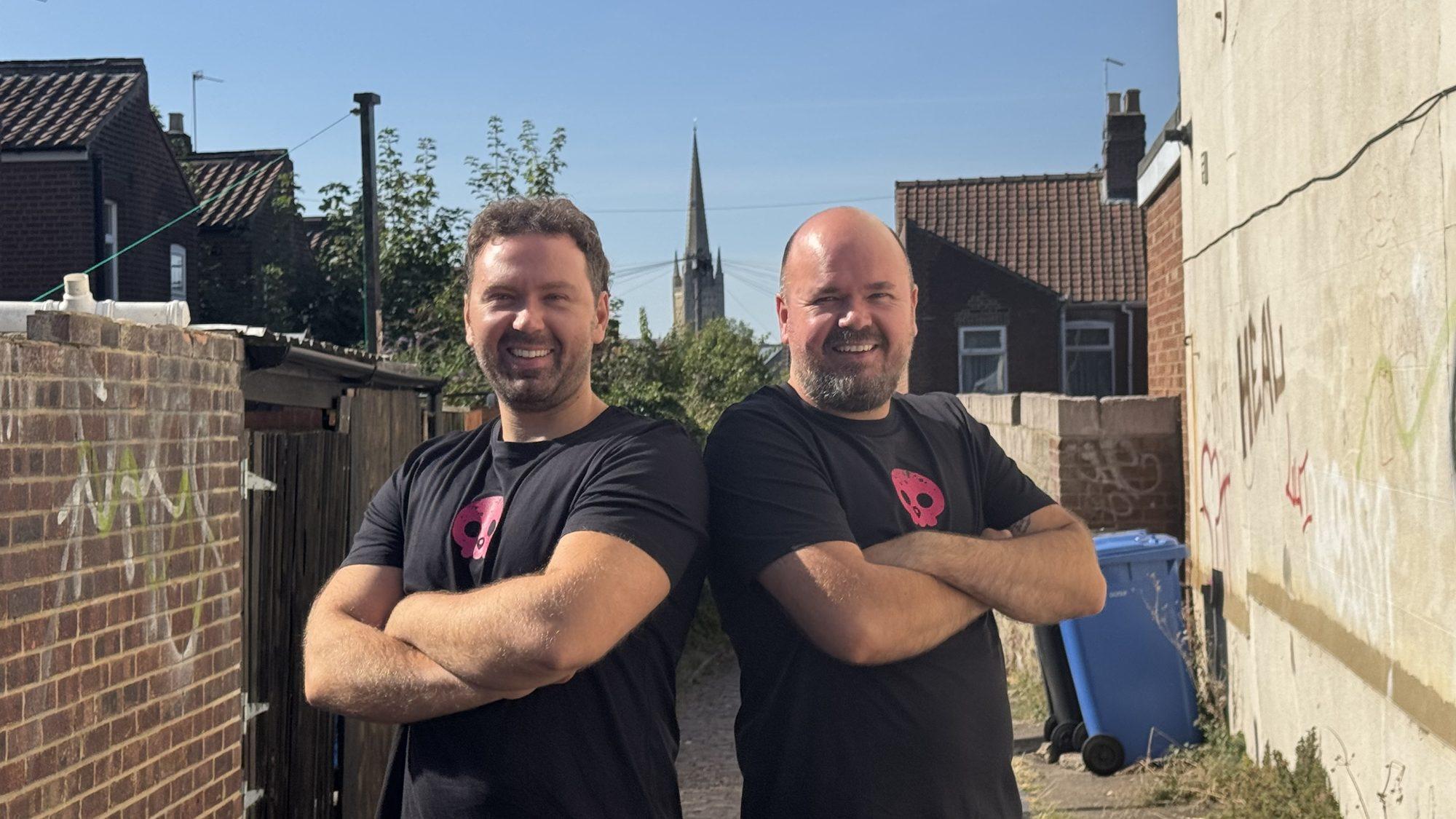 The two Area Autopsy co-founders standing arms-folded in a terraced back alley, with a cathedral spire behind them.