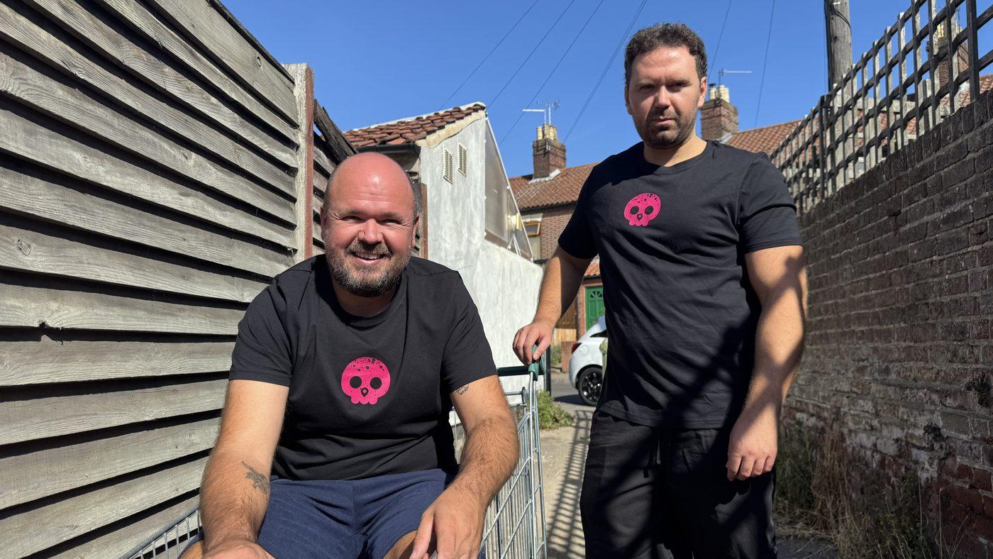 The two co-founders in an alleyway between terraced houses, one perched on an abandoned shopping trolley.